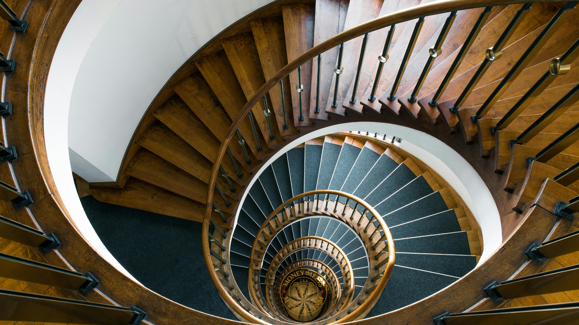An antique oak spiral staircase seen from above, winding down to the studio&rsquo;s seal at its centre.